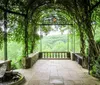 A serene garden pathway is flanked by lush greenery and vibrant flowers with a metal arch and a wooden bench inviting visitors to enjoy the surroundings
