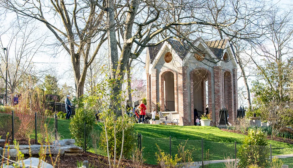 Visitors enjoy a sunny day at a park with a charming brick pavilion surrounded by budding trees and well-maintained gardens