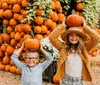 Two smiling children are playfully balancing pumpkins on their heads at a pumpkin patch adorned with numerous pumpkins and autumnal decorations in the background