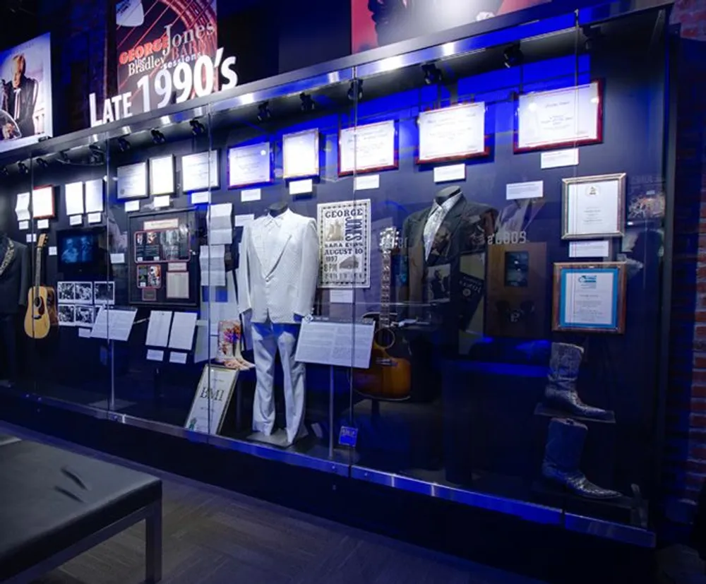 The image shows an exhibition case with various memorabilia including clothing guitars and framed documents likely from a music museum denoting a theme from the late 1990s