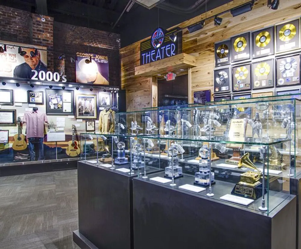 The image displays a music-themed exhibit featuring glass cases with awards and memorabilia guitars on display and decorated walls with images and gold records indicative of a musical museum or hall of fame