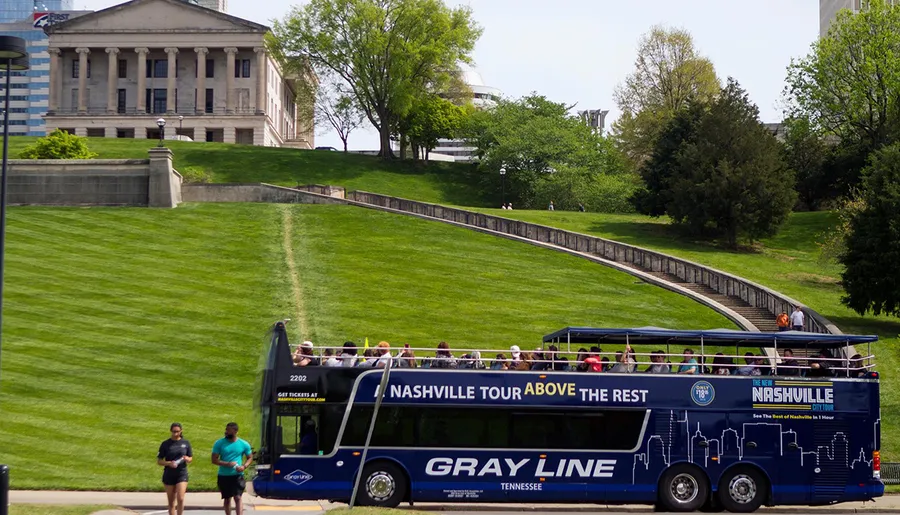 A double-decker tour bus labeled Gray Line with passengers on the open top is on a road with a grassy hill and a building atop in the background.