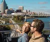 A couple is enjoying a scenic view from the top deck of a tour bus with a city skyline and a bridge in the background