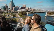 A couple is enjoying a scenic view from the top deck of a tour bus with a city skyline and a bridge in the background.