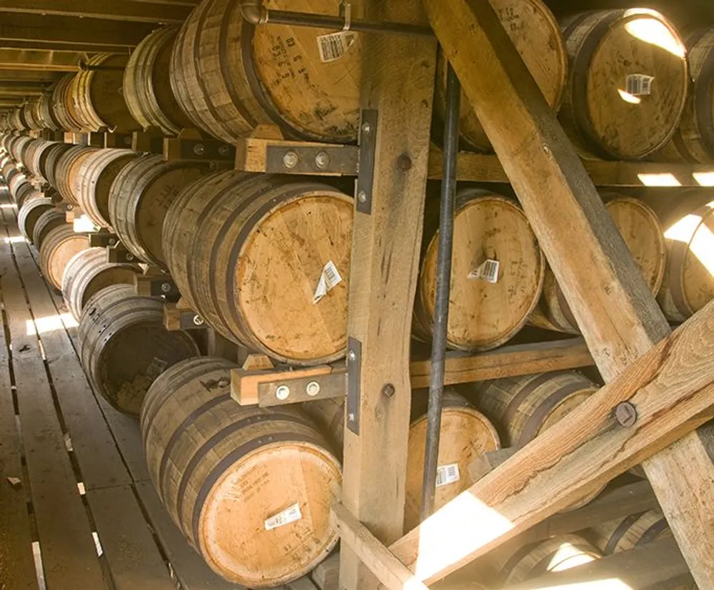 The image shows a collection of wooden barrels possibly containing whiskey or wine aging in a storage rack within a dimly lit warehouse