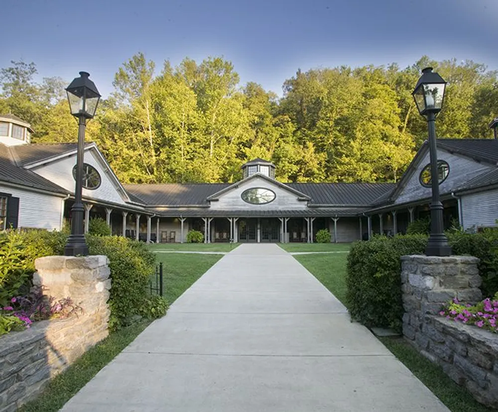 The image shows a symmetrical view of a stately building with a central clock feature flanked by two wings and fronted by a straight pathway traditional lamp posts and lush greenery giving off an air of serene elegance