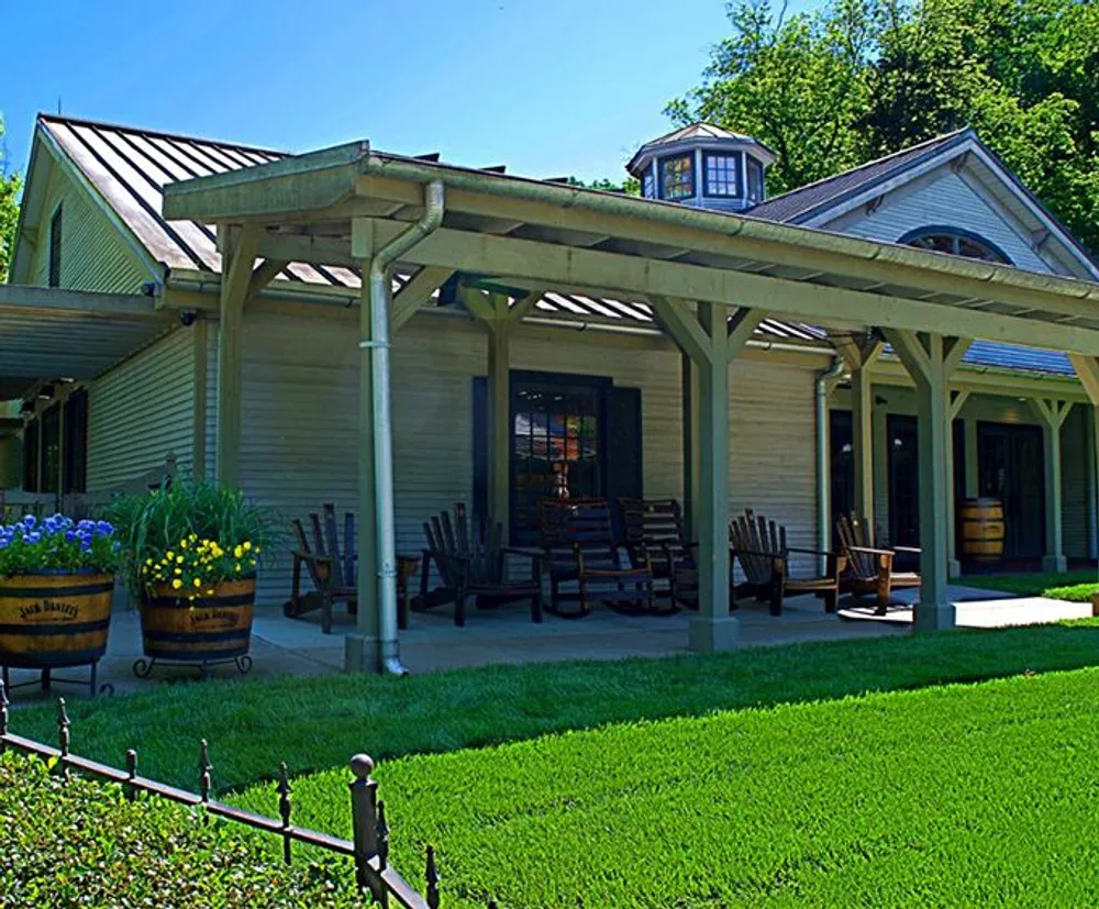The image shows a quaint wooden building with a covered porch area where several rocking chairs are lined up providing a relaxed seating area surrounded by greenery and adorned with flowering planters