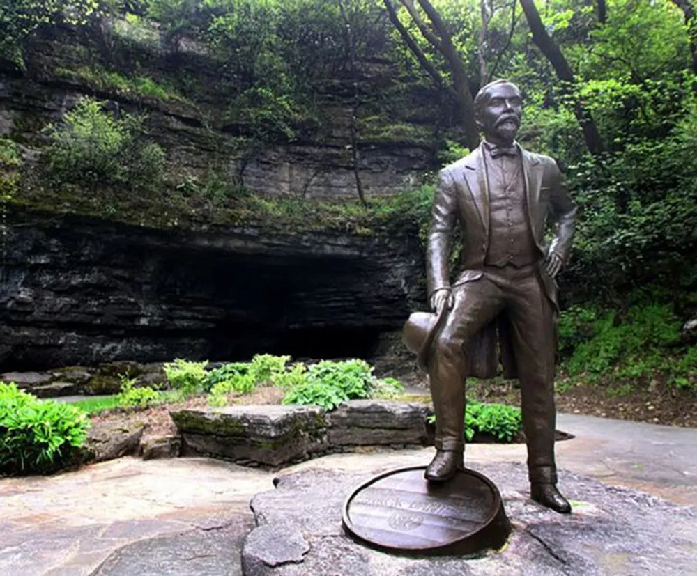 The image shows a bronze statue of a man standing confidently with a foot on a circular plate set against the backdrop of a lush green landscape with a cave entrance in the background