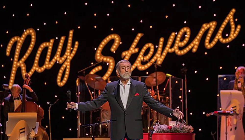 A man stands on stage with arms outstretched in front of a lit sign that displays the name Ray Stevens with musicians in the background