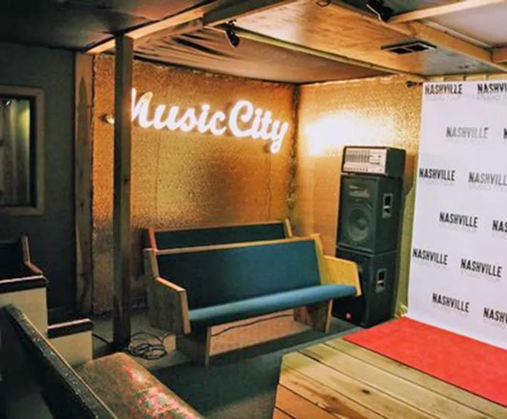 This image shows an indoor space with a neon sign that reads Music City a bench and a step-and-repeat banner with Nashville
