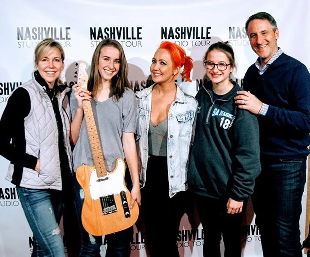 Five people are posing for a photo with smiles at a Nashville Studio Tour event where one person is holding a guitar
