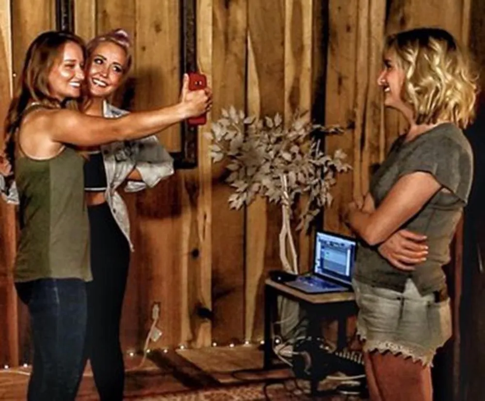Two women are taking a selfie while another woman watches them with a smile in a room with a rustic wooden wall and a laptop on a stand in the background