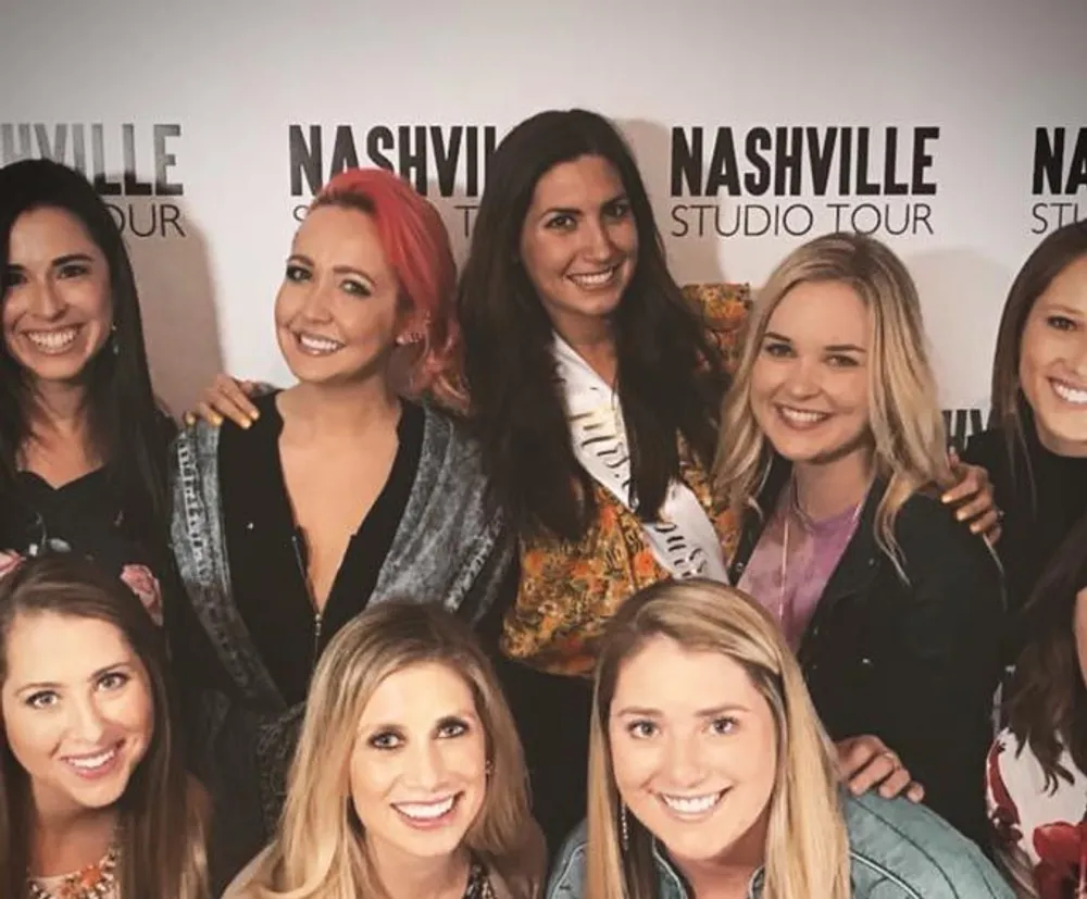 A group of smiling women pose together in front of a backdrop with the words Nashville Studio Tour