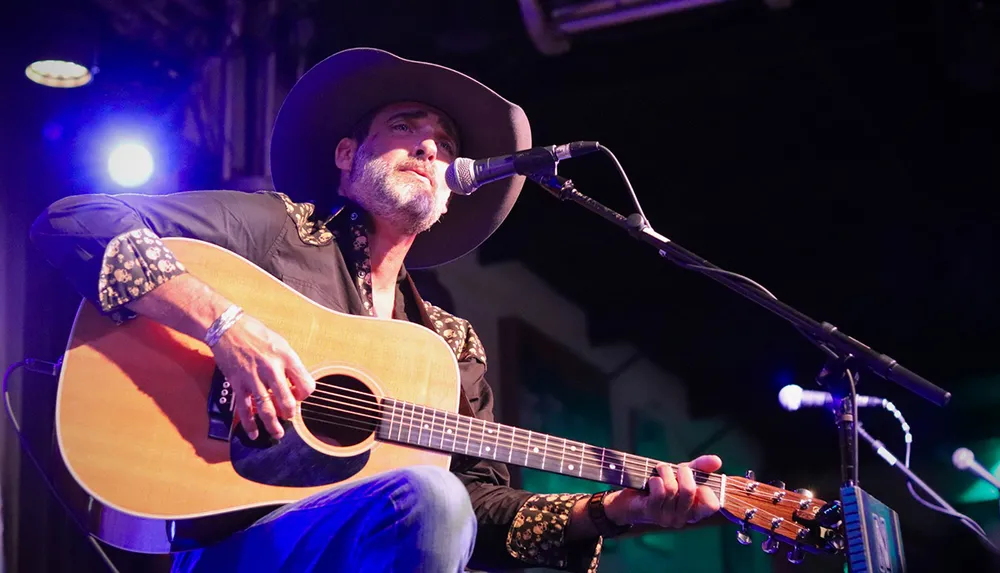 A musician in a cowboy hat is performing with an acoustic guitar on a stage singing into a microphone