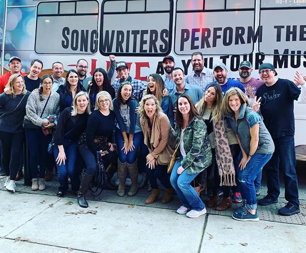 A group of cheerful people is posing for a photo in front of a bus that says Songwriters Perform Their Hit Songs Live On Tour