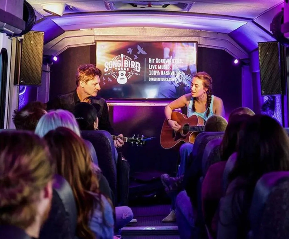A woman is playing a guitar and singing to an audience inside a bus adorned with music tour signage