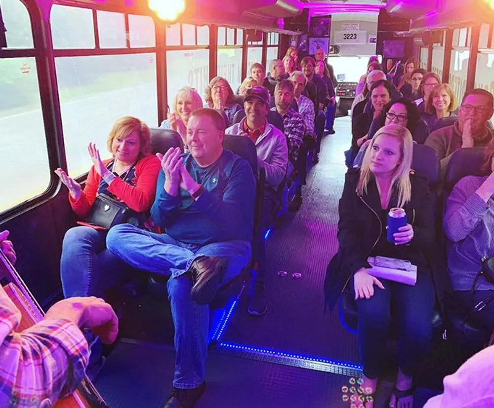 A group of people are sitting on a bus with purple lighting some smiling and clapping suggesting a lively atmosphere