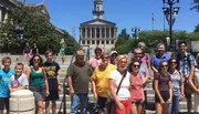 A diverse group of people are posing for a photo in front of a large building with a central dome on a sunny day.