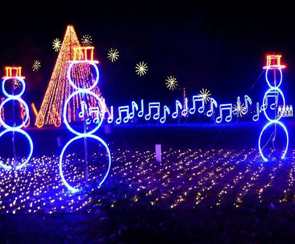 The image depicts a festive outdoor lights display featuring illuminated snowmen and musical notes with a carpet of twinkling lights covering the ground