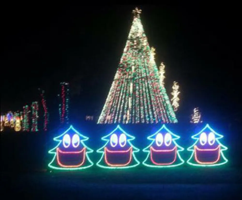 The image shows a festive outdoor display with a large illuminated Christmas tree and four smiley face decorations outlined in green and red lights in the foreground creating a cheerful nighttime holiday scene