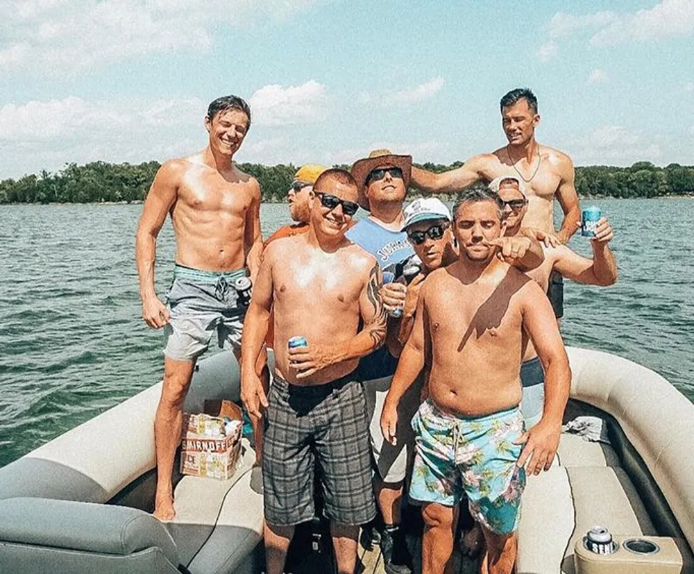 A group of cheerful men in swimwear are having a good time on a boat some holding beverages on a sunny day