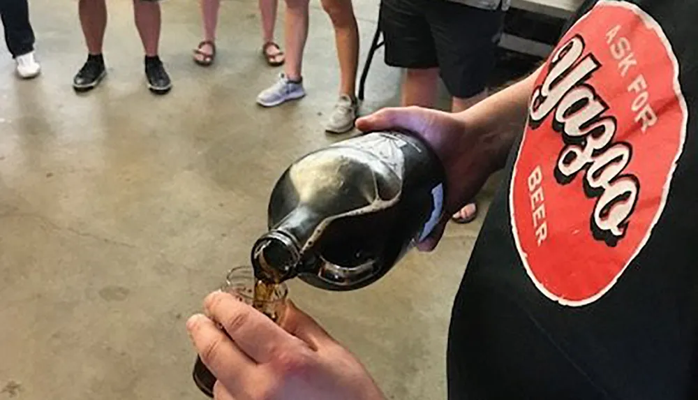 A person pours a dark liquid presumably beer from an oversized novelty bottle into a small clear cup while others watch
