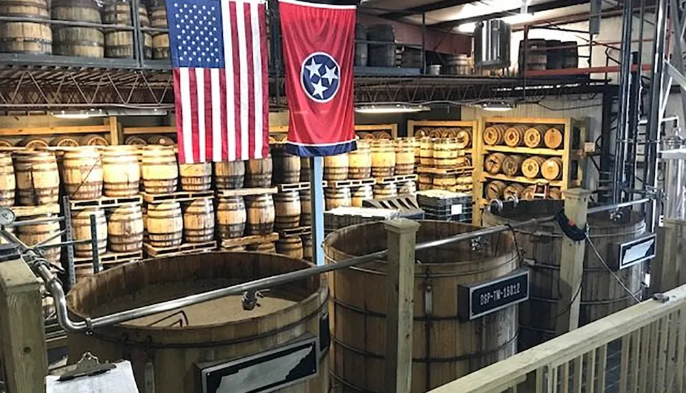 The image shows an interior view of a distillery with American flags and rows of wooden barrels potentially containing whiskey or another type of aged spirits