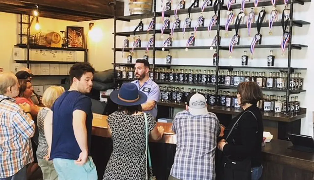 A group of customers is attentively listening to a staff member at a counter lined with jars and bottles likely at a food or beverage tasting event