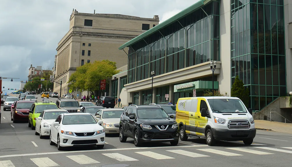 The image shows a busy urban street with mixed traffic including cars a taxi and a van with buildings and pedestrians on the sidewalk in the background