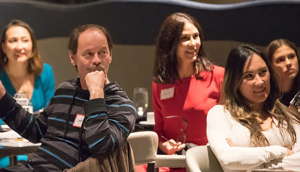 A group of people are seated and attentively engaged in a discussion or presentation with expressions ranging from thoughtful to smiling