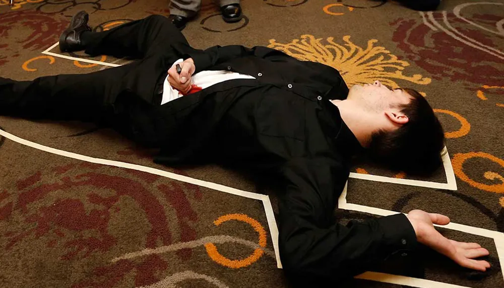 A man in a black suit is lying on the floor with his eyes closed holding a red object giving the appearance of an incident or a theatrical scene