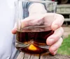 A person is holding a glass of amber liquid possibly whiskey with their right hand against a blurred outdoor background