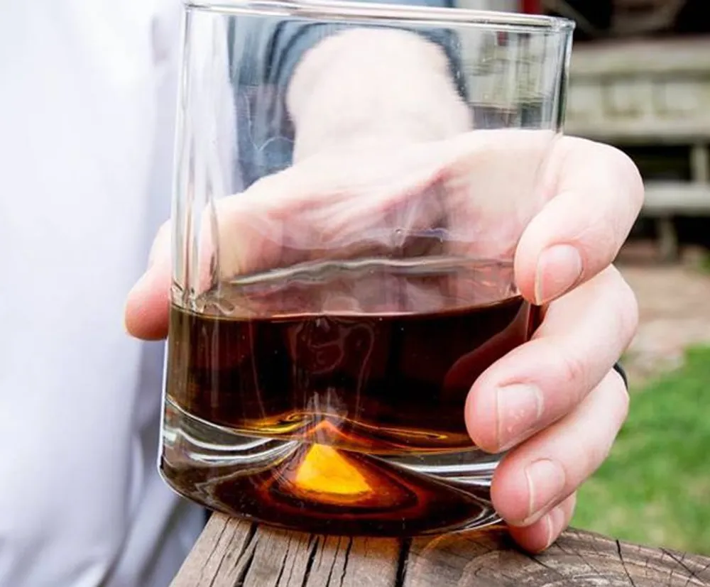 A person is holding a glass of amber liquid possibly whiskey with their right hand against a blurred outdoor background