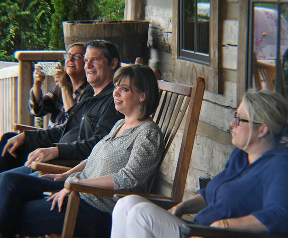 Four individuals are seated on wooden chairs on a porch with two appearing to enjoy a conversation one smiling towards the camera while holding a smartphone and the fourth looking away contemplatively
