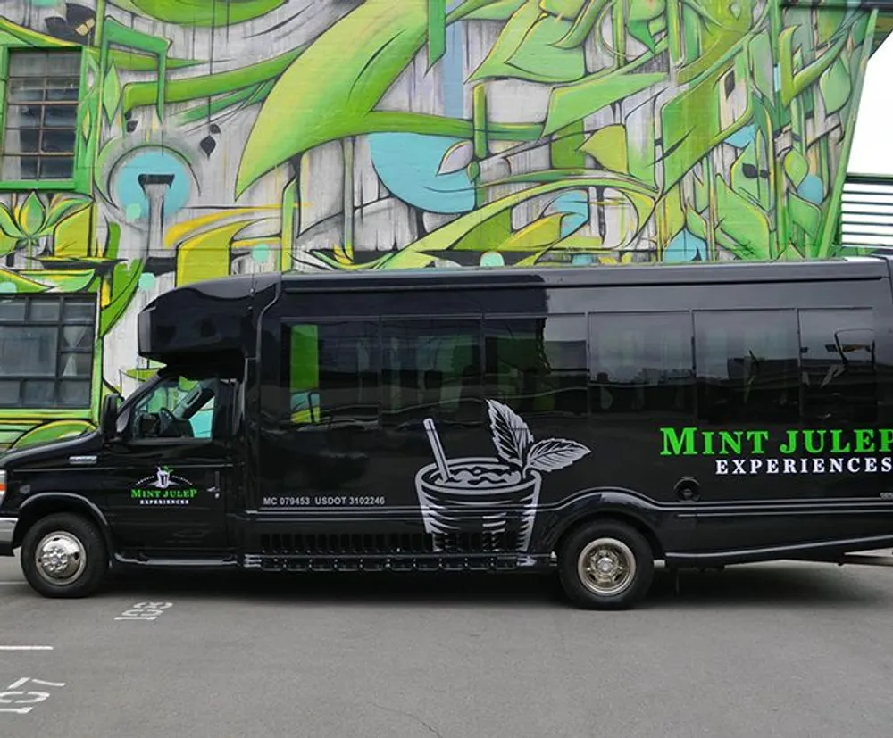 A black tour bus with Mint Julep Experiences on the side is parked in front of a large vibrant abstract graffiti mural