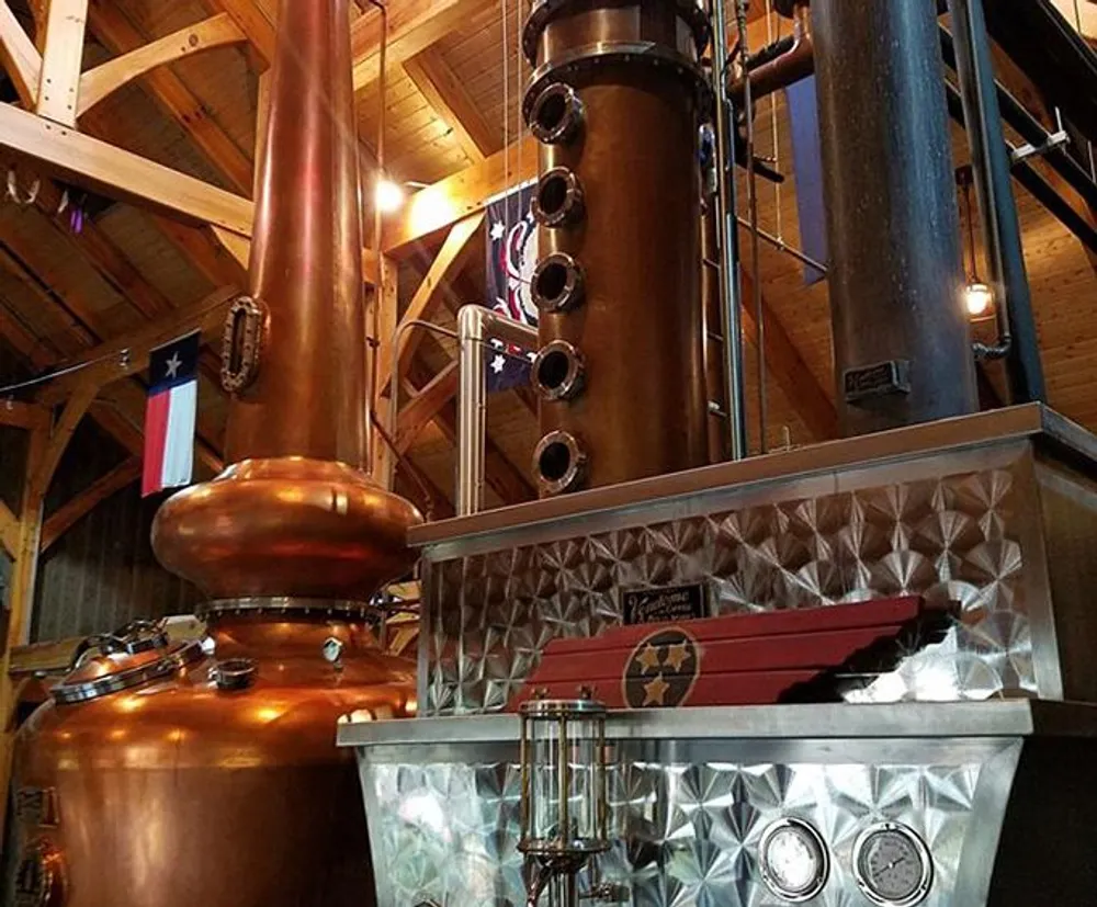 This image shows a copper distillation apparatus possibly in a whiskey or spirits distillery with an American flag and another flag featuring a star reflecting a patriotic or Texan theme within a wooden-beamed structure