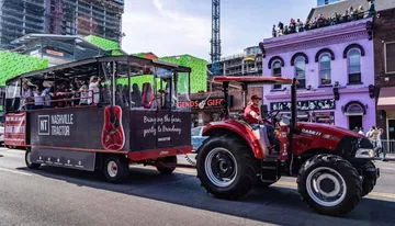 A tractor is pulling a party trailer filled with people on a sunny day in an urban environment.