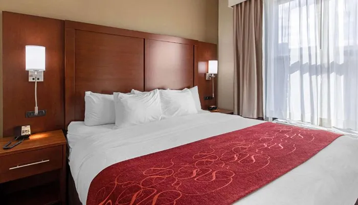 The image shows a neatly made hotel bed with white linen and a red decorative bed runner flanked by wooden nightstands and wall-mounted lamps against a backdrop of a curtained window and a wooden headboard