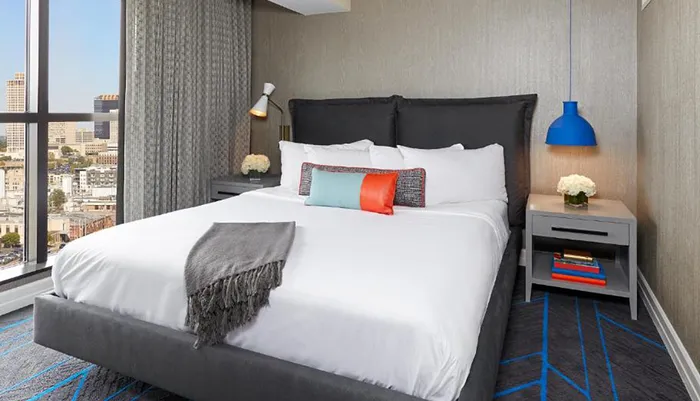The image shows a modern hotel room with a large bed colorful accents and a view of an urban landscape outside the window