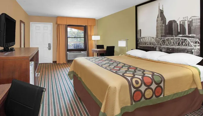 The image shows a neatly organized hotel room with a patterned bedspread a work desk a mounted flat-screen TV and a large wall art of a city skyline