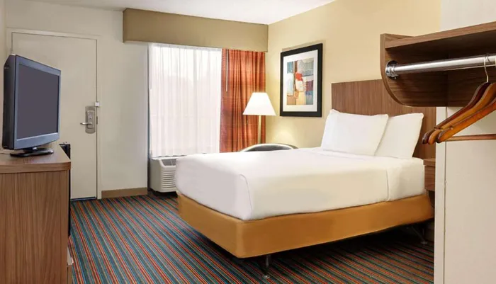 The image shows a neatly arranged hotel room with a bed a television set a window with curtains and decorative elements such as a picture frame and carpeting