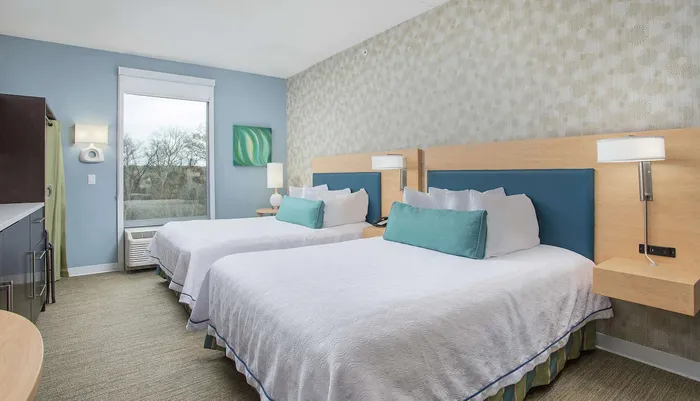 This image features a well-lit tidy hotel room with two queen-sized beds decorated in calming blue and green tones and a view of trees through the window