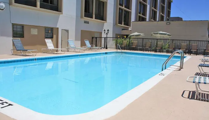 The image shows a sunny outdoor swimming pool area with loungers and umbrellas surrounded by a building with multiple floors