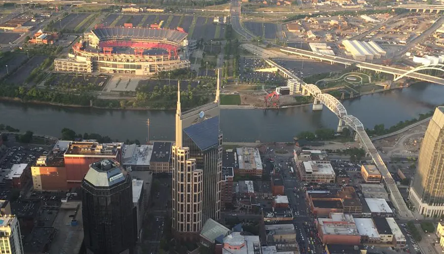 This image shows an aerial view of a cityscape with a stadium on the left, a river flowing through the middle, various buildings, and a distinctive bridge crossing the river.