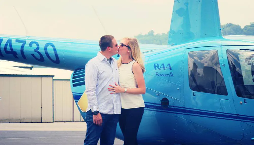 A couple is sharing a kiss in front of a blue R44 Raven II helicopter