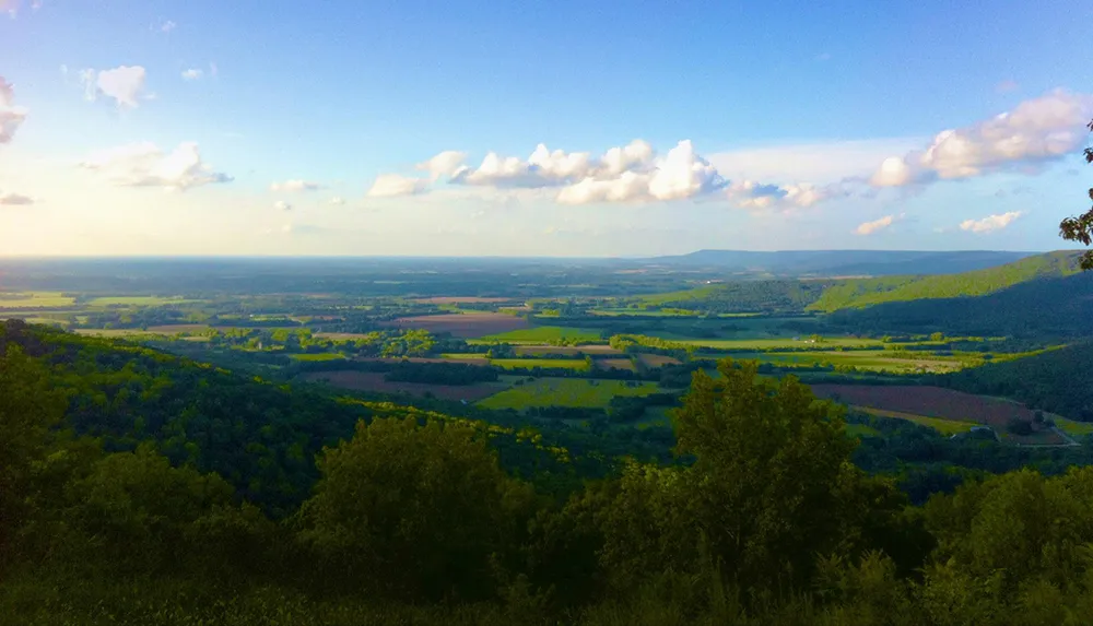 The image displays a scenic view of a lush valley with patchwork fields and forests stretching towards distant hills under a sky dotted with clouds