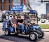 Four individuals are seated in an open-sided branded golf cart-like vehicle advertising tours and beer with an urban street scene in the background