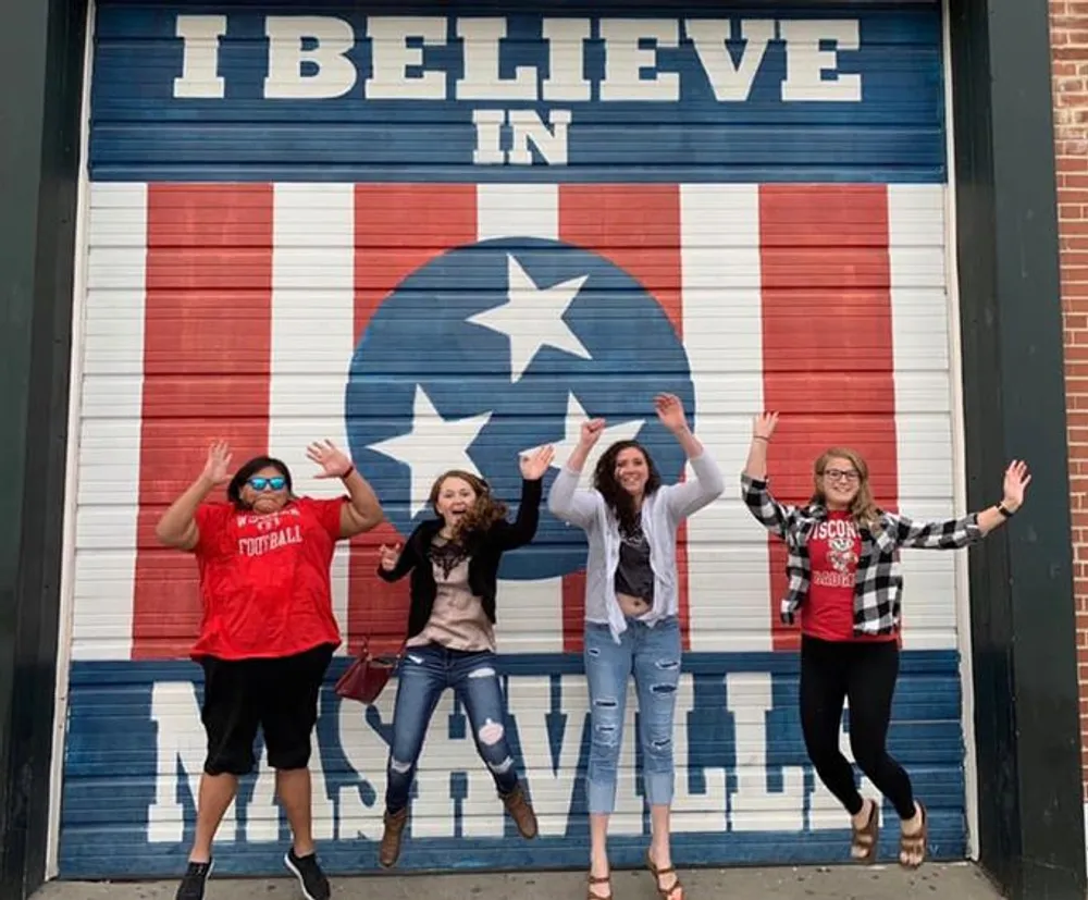 Four joyful people are posing with raised arms in front of a mural that reads I BELIEVE IN NASHVILLE which has the American flags colors and a large central star