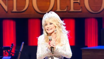 The image shows a performer singing into a microphone on stage, with a lit sign in the background and musical instruments visible in the foreground.