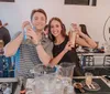 Two people are smiling at the camera holding cocktail shakers with various bar equipment and glasses on the table in front of them suggesting they are participating in a cocktail-making class or event